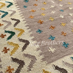 May include: Close-up of a woven rug with a geometric pattern. The rug features a cream-colored border with brown, yellow, and teal arrow-like designs. The central field is a muted mauve color with scattered floral motifs in various colors. The text "Rug Brothers" is visible.