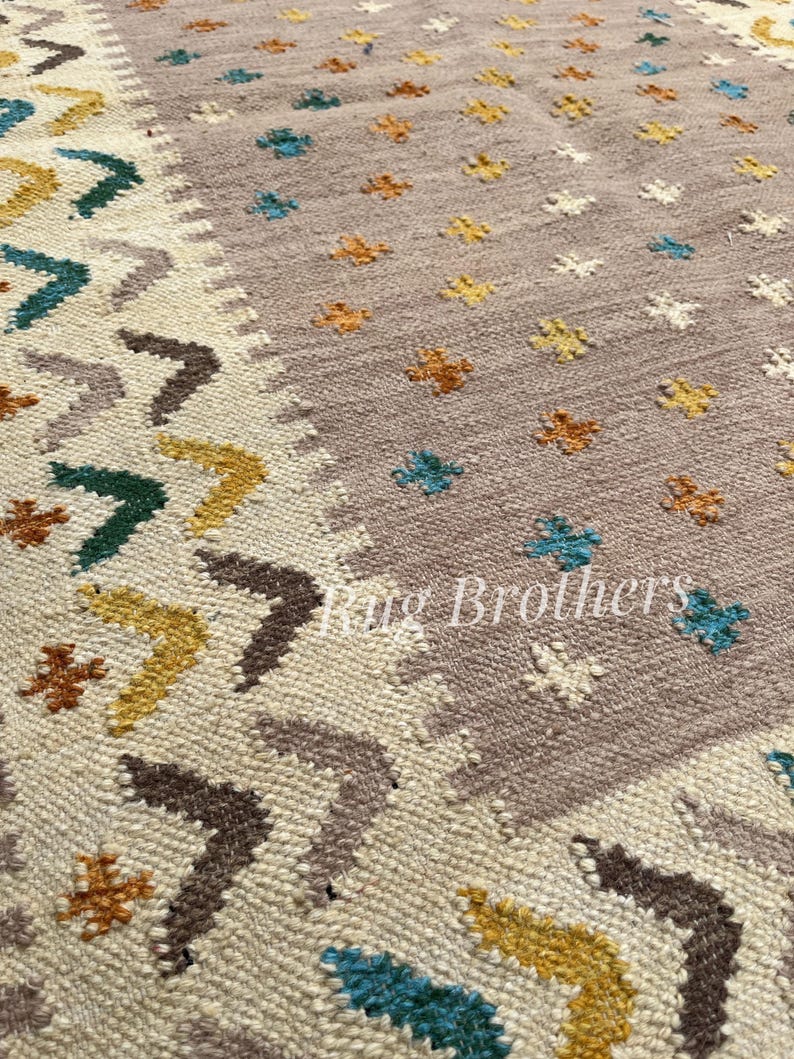 May include: Close-up of a woven rug with a beige and mauve background. The rug features a repeating pattern of arrows and floral motifs in shades of brown, yellow, teal, and orange. The text "Rug Brothers" is visible.