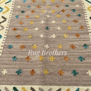 May include: A rectangular rug with a central field of taupe, adorned with small, colorful floral patterns in yellow, teal, and brown. The border features geometric designs in cream, brown, teal, and yellow. The text "Rug Brothers" is in the center.