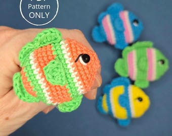 May include: A hand holding a crocheted orange, green, and white fish with a black eye. Three other crocheted fish in blue, green, and yellow with pink and white stripes are in the background. The image includes a circle with the text "PDF Pattern ONLY".