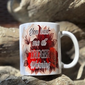 Può includere: Tazza in ceramica bianca con la scritta "You had me at TRIGGER Warning" in un carattere decorativo. La tazza presenta un design floreale con rose rosse e gigli bianchi. La tazza ha un manico bianco.