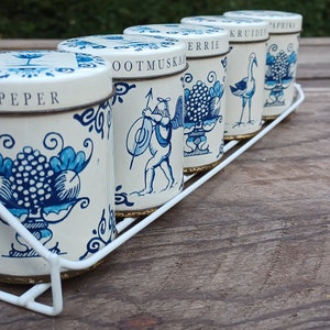 May include: Five white ceramic spice jars with blue and gold designs. The jars are labeled "Pepper", "Ootmus", "Errie", "Kruiden", and "Paprika". The jars are on a white metal rack.