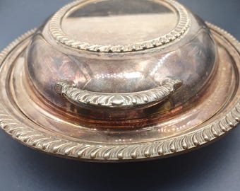 Può includere: Un piatto da portata in argento con coperchio e manico decorativo. Il piatto ha un bordo rialzato e una superficie liscia e lucida.
