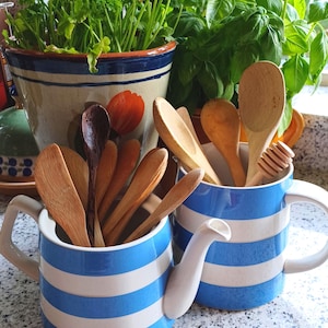 May include: Two blue and white striped ceramic teapots filled with wooden spoons. One teapot has a spout and handle. Herbs and plants are in the background, including parsley and basil. A honey dipper is visible.