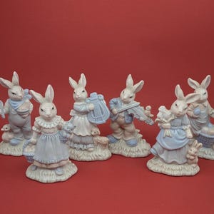 Può includere: Una collezione di statuine di conigli in ceramica in varie pose, vestite con abiti azzurro pastello e bianchi. Alcuni conigli tengono strumenti musicali, cesti o piccoli animali. Le statuine sono ambientate su uno sfondo rosso.