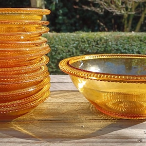 May include: A stack of amber-colored glass plates and a matching bowl. The plates and bowl have a beaded rim design. The glass is transparent, allowing light to pass through, and is sitting on a wooden surface.