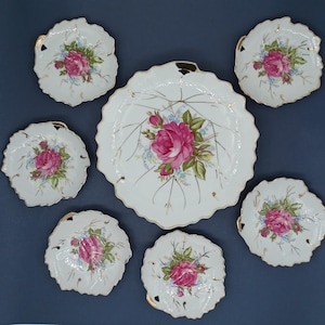 Può includere: Set di sette piatti in porcellana bianca con bordi dorati e motivi floreali. I piatti presentano rose rosa e foglie verdi su uno sfondo bianco. I piatti hanno la forma di foglie e hanno un bordo ondulato.