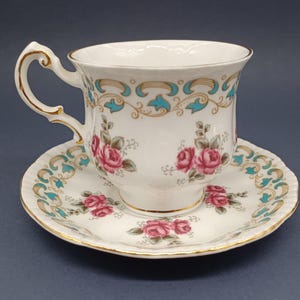 May include: A white teacup and saucer set with a floral design. The cup and saucer feature a delicate pattern of pink roses and green leaves, accented with gold trim and a turquoise blue border.
