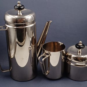 Può includere: Un set da caffè in argento composto da caffettiera, lattiera e zuccheriera. La caffettiera ha un beccuccio e un manico. La lattiera e la zuccheriera hanno coperchi con pomelli neri.