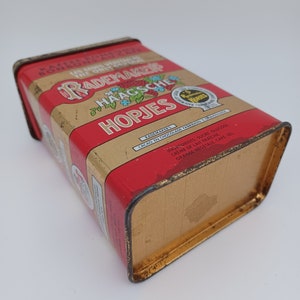 May include: A red and gold tin box with the text "Rademaker Haagsche Hopjes" on the front. The text "Rademaker" is in a stylized font and the text "Haagsche Hopjes" is in a smaller font. The tin box is slightly worn and has a few scratches.