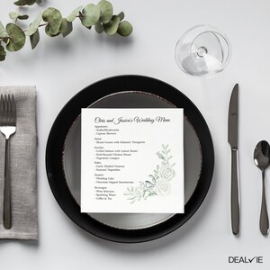 May include: A square wedding menu card featuring the text Chris and Jessica's Wedding Menu with a list of food items and a watercolor illustration of green leaves and white roses in the bottom right corner, placed on a black and gray plate.