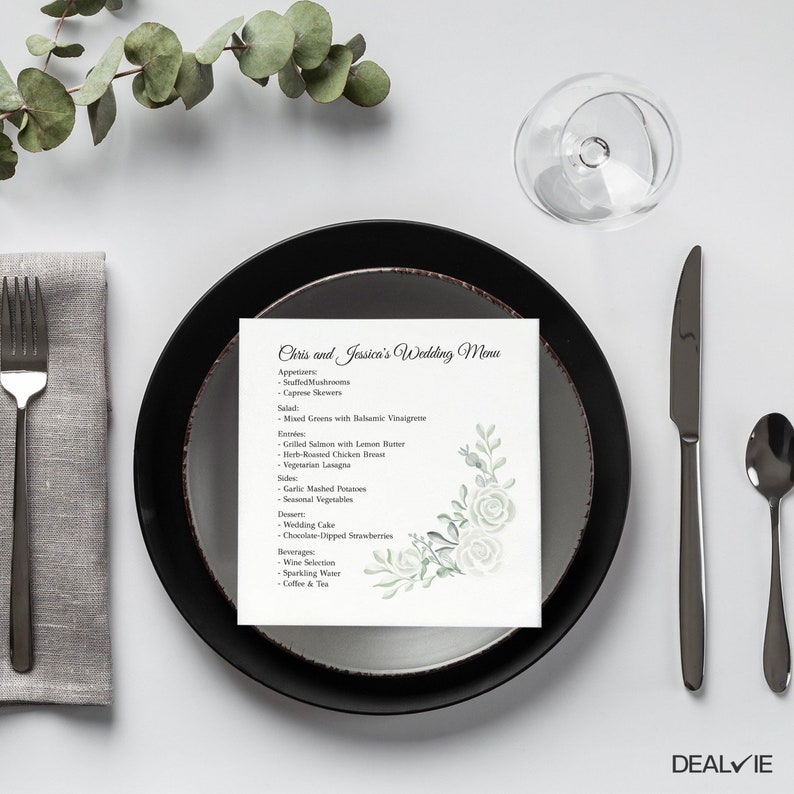 May include: A square wedding menu card featuring the text Chris and Jessica's Wedding Menu with a list of food items and a watercolor illustration of green leaves and white roses in the bottom right corner, placed on a black and gray plate.