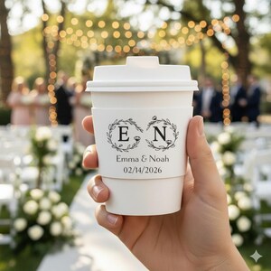 May include: A white paper coffee cup with a lid and a decorative sleeve. The sleeve features the initials "E" and "N" inside laurel wreaths, along with the names "Emma & Noah" and the date "02/14/2026". The cup is held in front of a blurred outdoor wedding scene.