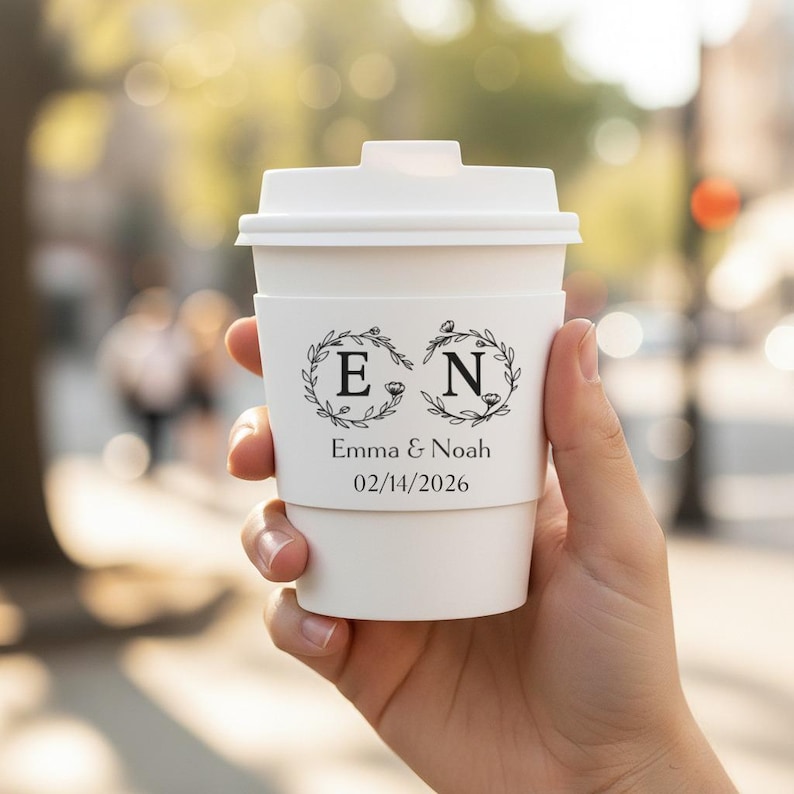 May include: A white paper coffee cup with a lid and a sleeve. The cup features the initials "E" and "N" inside floral wreaths, along with the names "Emma & Noah" and the date "02/14/2026".