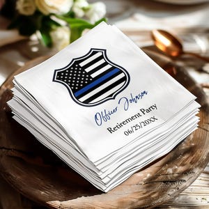 May include: A stack of white paper napkins featuring a black and white police shield graphic with a thin blue line. The text reads Officer Johnson, Retirement Party, and 06/25/20XX in a combination of blue script and black serif fonts.