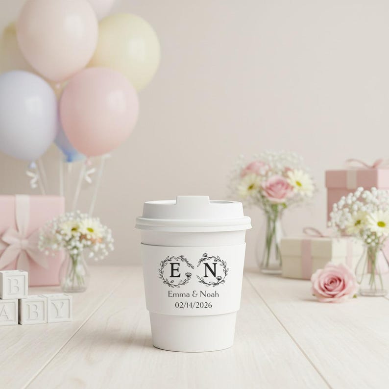 May include: A white paper coffee cup with a lid, featuring a floral design with the letters "E" and "N" and the date 02/14/2026. The cup is surrounded by balloons, flowers, and gift boxes, suggesting a celebration or event.