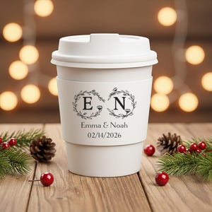 May include: A white travel coffee mug with a lid and a decorative band. The band features the initials "E" and "N" within floral wreaths, along with the names "Emma & Noah" and the date "02/14/2026". The mug is surrounded by pinecones and berries.