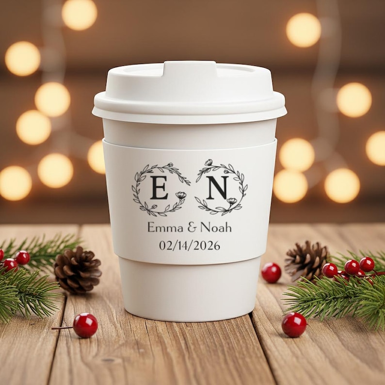 May include: A white travel coffee mug with a lid and a decorative band. The band features the initials "E" and "N" within floral wreaths, along with the names "Emma & Noah" and the date "02/14/2026". The mug is surrounded by pinecones and berries.