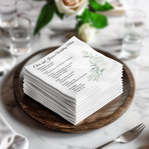 May include: A stack of white square paper napkins featuring the text Chris and Jessica's Wedding Menu in black script and a printed illustration of pale green leaves and white roses on the right side, resting on a dark brown wooden plate.