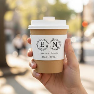May include: A disposable coffee cup with a white lid and a custom sleeve. The sleeve features the initials "E" and "N" inside laurel wreaths, along with the names "Emma & Noah" and the date "02/14/2026".