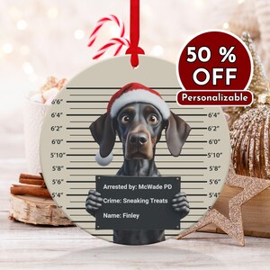 Puede incluir: Adorno navideño redondo con un perro con gorro de Papá Noel, posando para una foto policial. El perro sostiene un letrero que dice "Arrestado por: McWade PD, Delito: Sneaking Treats, Nombre: Finley". El adorno tiene una cinta roja y una pegatina "50% OFF".