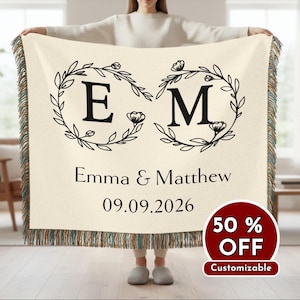 May include: Cream-coloured personalised throw blanket with black floral wreath design, initials "E" and "M", names "Emma & Matthew", and date "09.09.2026". The blanket has a fringed edge.