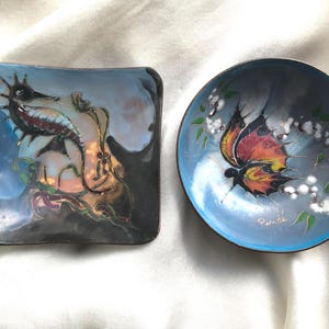 Może przedstawiać: Dwa ozdobne talerze emaliowane z niebieskim tłem. Jeden talerz przedstawia konika morskiego z niebieskim, czerwonym i złotym wzorem. Drugi talerz przedstawia motyla z czerwonymi, żółtymi i czarnymi skrzydłami oraz tekstem "Paris de".