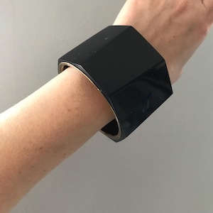 Könnte beinhalten: Ein schwarzes achteckiges Armreif-Armband mit einem goldenen Metallband.