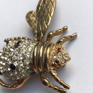 May include: A gold-toned bee brooch with a textured wing and a body covered in clear and black rhinestones. The bee has red rhinestone eyes.