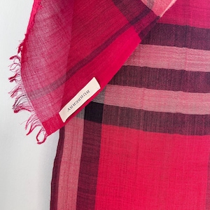 May include: A vibrant red scarf with a classic plaid pattern. The scarf features a fringed edge and a small white tag with the brand name "BURBERRY". The design incorporates shades of red and black, creating a bold and stylish accessory.