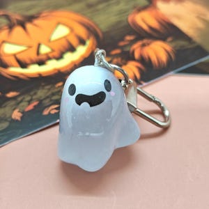 May include: A light blue ghost-shaped keychain with a silver clasp. The ghost has black eyes, a black mouth, and pink cheeks. The background includes a Halloween-themed illustration with a jack-o'-lantern.