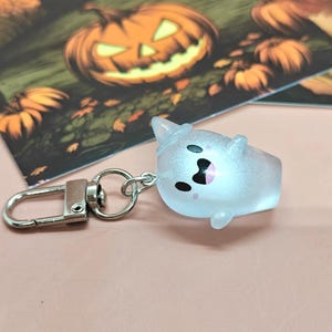 May include: A light blue ghost-shaped keychain with a silver clasp. The ghost has black eyes and a pink mouth, and is wearing a witch hat. The background features a Halloween-themed illustration with pumpkins.