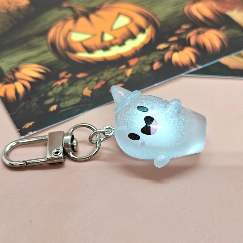 May include: A light blue ghost-shaped keychain with a silver clasp. The ghost has black eyes and a pink mouth, and is wearing a witch hat. The background features a Halloween-themed illustration with pumpkins.