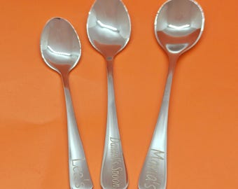 May include: Three stainless steel spoons with engraved names: "Lea's", "Daniel's Spoon", and "Maria's". The spoons are arranged vertically against an orange background. The spoons have a reflective, polished surface.