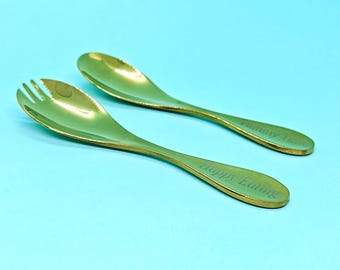 May include: Two gold-coloured serving utensils on a light blue background. One is a spoon, the other a fork-like spoon. The text "Happy Eating" and "Yummy Taste" are engraved on the handles.