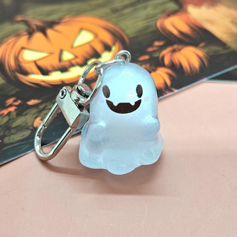 May include: A translucent ghost-shaped keychain with a silver clasp. The ghost has a smiling face with black eyes and a brown mouth. The background includes a blurred image of a jack-o'-lantern, suggesting a Halloween theme.