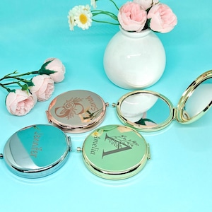 Peut inclure: Plusieurs miroirs de poche personnalisés en argent, or rose et vert. Chaque miroir présente un design unique avec des noms ou des initiales et des motifs floraux. Les miroirs sont ouverts et fermés, révélant leurs surfaces réfléchissantes. Un vase de fleurs est en arrière-plan.