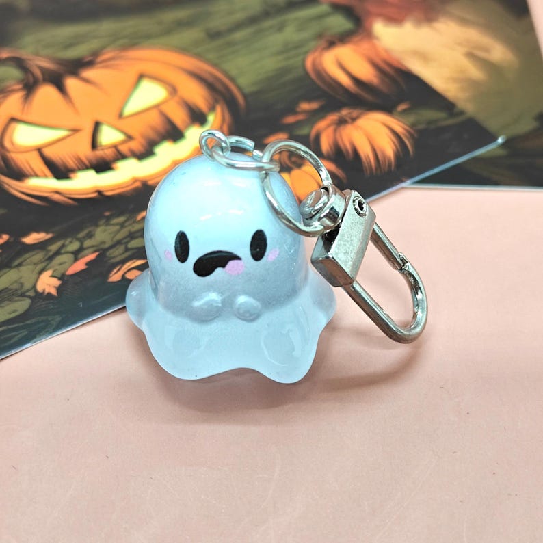 May include: A light blue ghost-shaped keychain with a silver clasp. The ghost has black eyes, a black mouth, and pink cheeks. The background features a Halloween-themed illustration with a jack-o'-lantern.