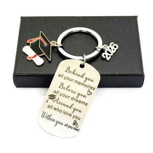 May include: A silver keychain with a graduation cap charm, a diploma charm, and a 2026 charm. The dog tag-shaped pendant has an engraved message: "Behind you all your memories, Before you all your dreams, Around you all who love you, Within you all you need."