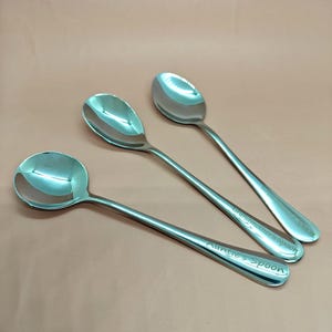 May include: Three stainless steel spoons with engraved text. One spoon reads "Auntie's Spoon", and another reads "Grandmother's Spoon". The spoons have a polished, reflective surface and are arranged on a light brown surface.
