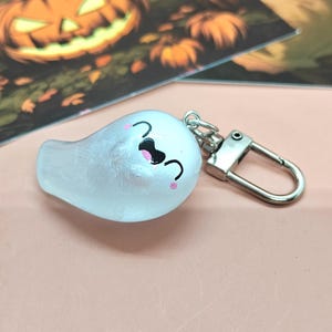 May include: A white ghost-shaped keychain with a smiling face, black eyes, and pink cheeks. The keychain has a silver clasp and is likely a Halloween-themed accessory.
