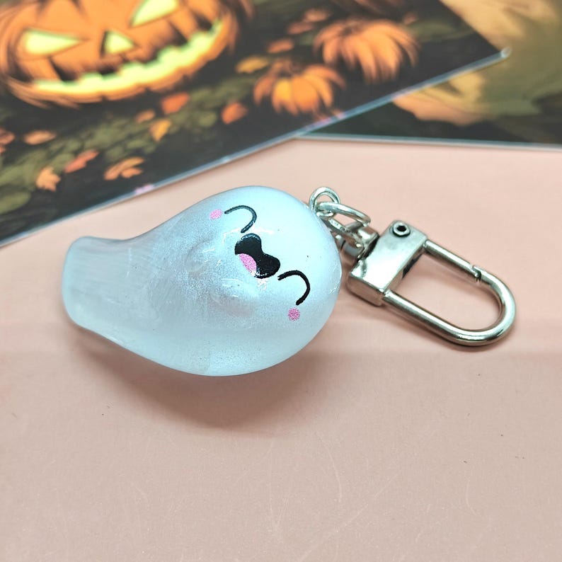 May include: A white ghost-shaped keychain with a smiling face, black eyes, and pink cheeks. The keychain has a silver clasp and is likely a Halloween-themed accessory.