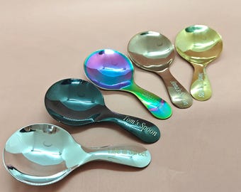 May include: A collection of five metal spoons with rounded bowls and engraved handles. The spoons have various finishes, including silver, iridescent, and gold. The handles are engraved with phrases like "Tom's Spoon" and "Life is Sweet."