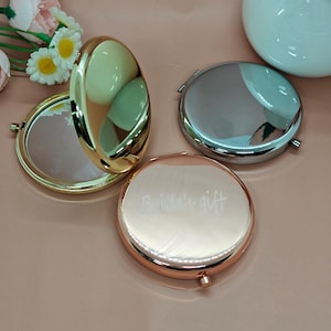 以下が含まれることがあります： 金、銀、ローズゴールドの丸いコンパクトミラーのコレクション。1つは開いており、鏡面が見えます。ローズゴールドのミラーには「Bride's gift」の文字が刻まれています。ミラーは丸く、光沢のある仕上がりです。