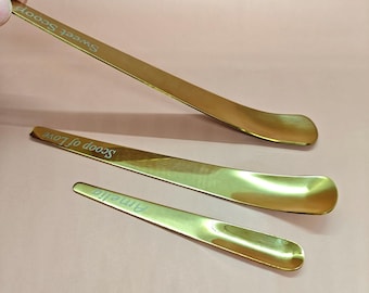 May include: Three gold-coloured serving spoons with curved, flat scoop ends. The spoons have text engraved on the handles: "Sweet Scoop", "Scoop of Love", and "Amelia". The spoons are arranged on a pink surface.
