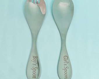 May include: Two silver serving spoons, one with a fork-like end and the other a solid spoon. The spoon with the fork end is labeled "Boy's Spoon", and the other is labeled "Girl's Spoon". The background is a light blue.