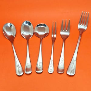 May include: Six stainless steel spoons and forks are arranged on an orange background. Each piece is engraved with a name or phrase, including "Wedding Gift," "Jack's Spoon," and "Tom's Gift." The utensils are for dining.