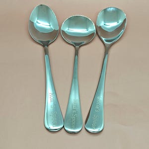 Puede incluir: Tres cucharas de acero inoxidable pulido con texto grabado. Las cucharas están dispuestas una al lado de la otra. El texto de las cucharas dice "Mom's Spoon", "Dad's Spoon" y "Uncle's Spoon".
