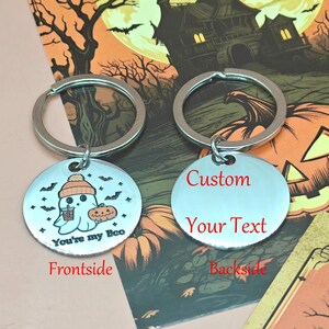 May include: Two silver Halloween keychains. One keychain features a ghost with a pumpkin and the text "You're my Boo". The other keychain has the text "Custom Your Text". The keychains are on a Halloween-themed background.