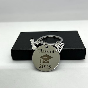 Puede incluir: Llavero plateado con un dije de gorro de graduación, un dije de rollo y una etiqueta redonda que dice "Clase de 2025".